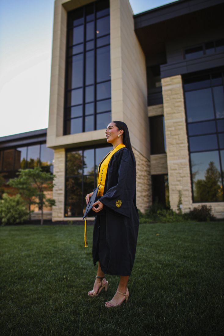 A graduate in a yellow stole standing on the lawn beside a modern campus building