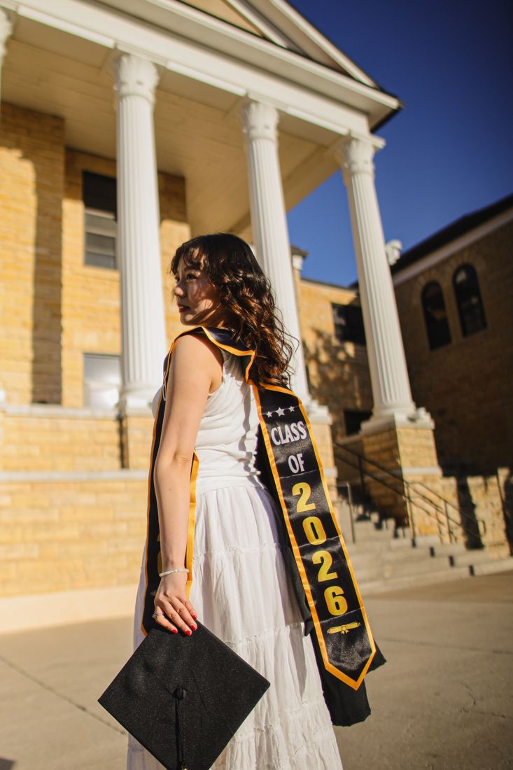 A graduate in a white dress with a Class of 2026 sash, looking away from the camera in front of a columned building